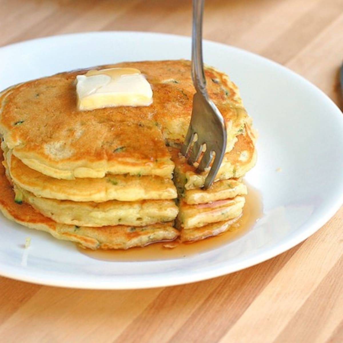 Stack of zucchini pancakes with butter and maple syrup with a fork.